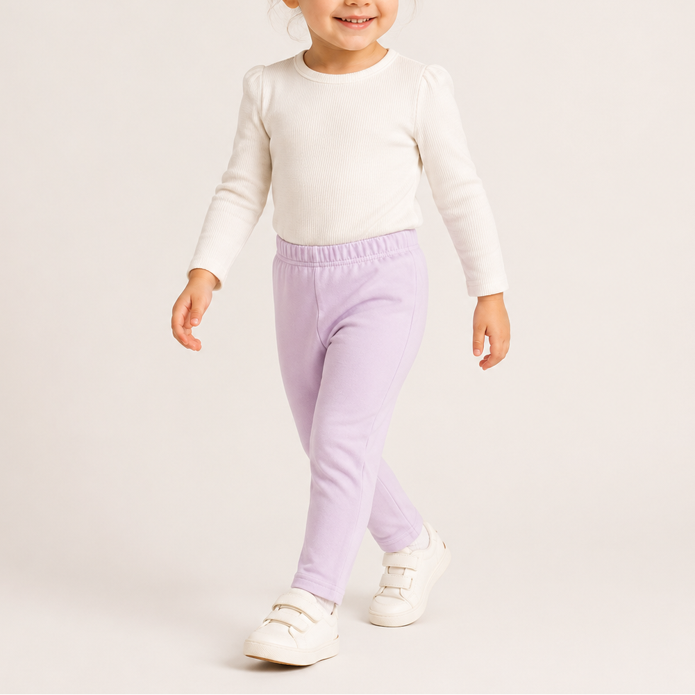 A toddler girl walking and smiling, wearing a cream long-sleeve knit top, light purple elastic-waist pants, and white velcro sneakers against a plain light beige studio background.