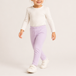 A toddler girl walking and smiling, wearing a cream long-sleeve knit top, light purple elastic-waist pants, and white velcro sneakers against a plain light beige studio background.