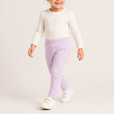 A toddler girl walking and smiling, wearing a cream long-sleeve knit top, light purple elastic-waist pants, and white velcro sneakers against a plain light beige studio background.