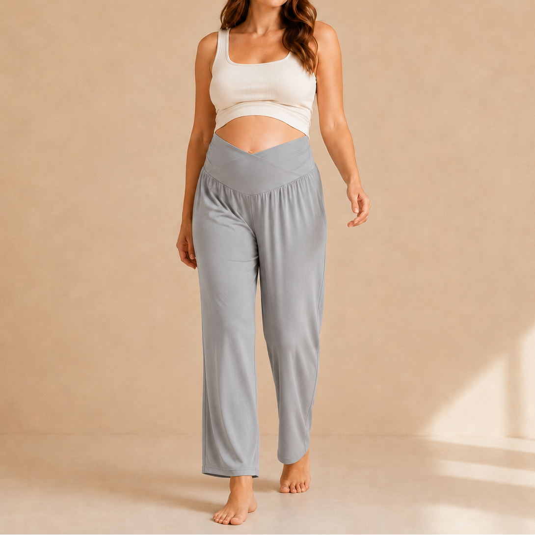 A woman stands barefoot in a studio wearing light blue high-waisted lounge pants and a white cropped tank top against a warm beige backdrop.