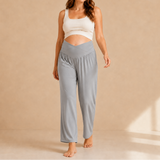 A woman stands barefoot in a studio wearing light blue high-waisted lounge pants and a white cropped tank top against a warm beige backdrop.