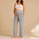 A pregnant woman stands barefoot in a studio wearing light gray maternity pants and a white crop top, with her hands on her belly, against a warm beige background.
