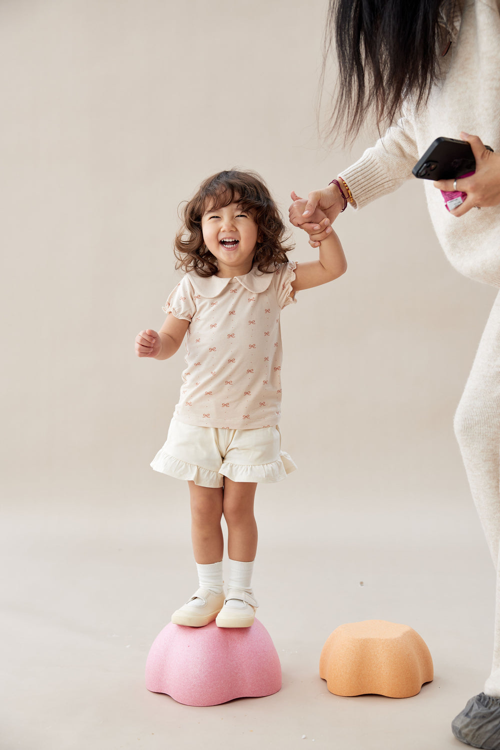 Child standing on a pink balance toy with an adult holding their hand, on a plain background