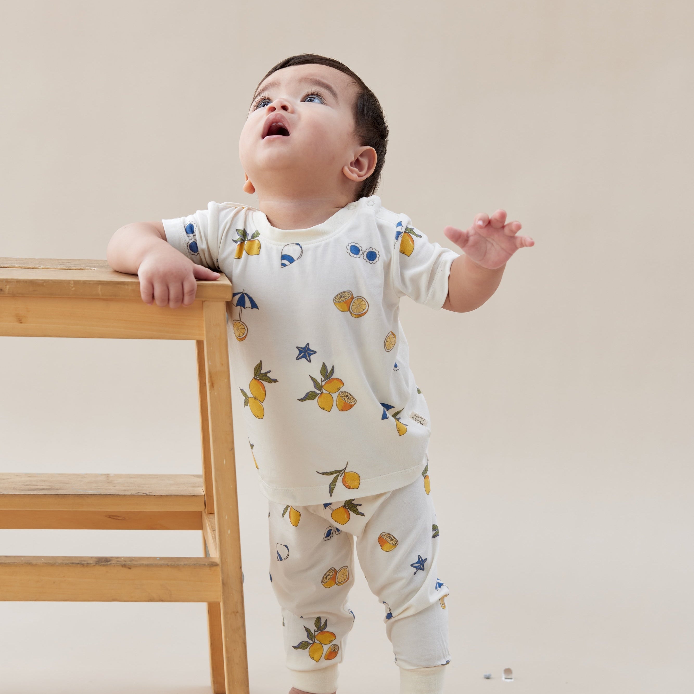 Baby in a lemon home set standing next to a wooden stool against a plain background
