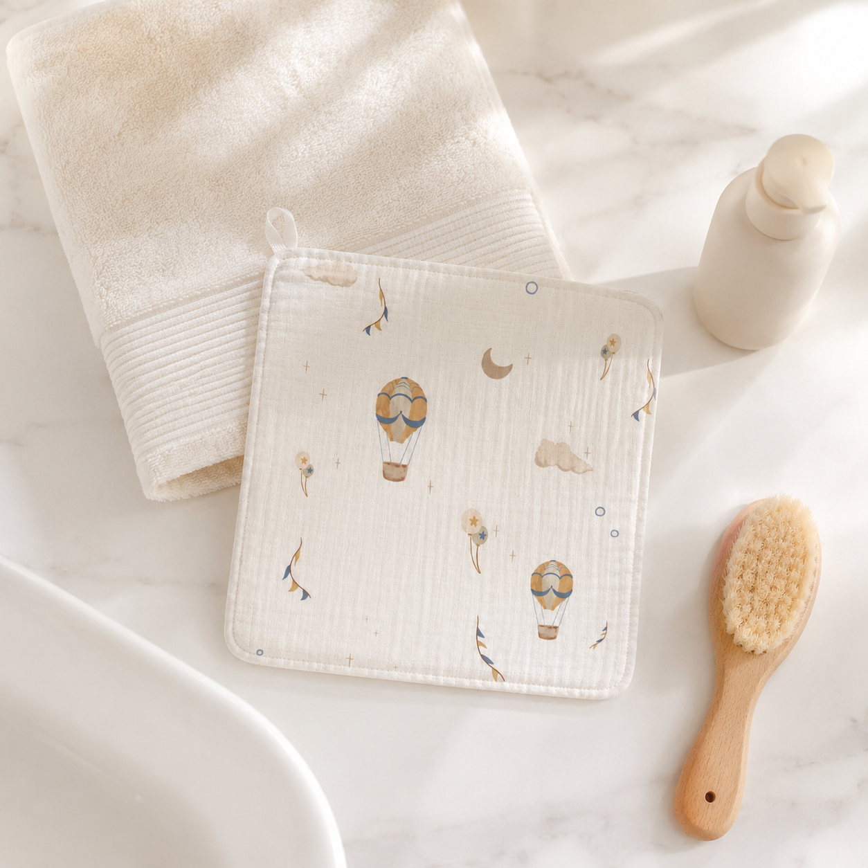 A square muslin baby towel or cloth with a hot air balloon print, styled on a white marble surface next to a folded cream towel, a small bottle, and a wooden baby brush.