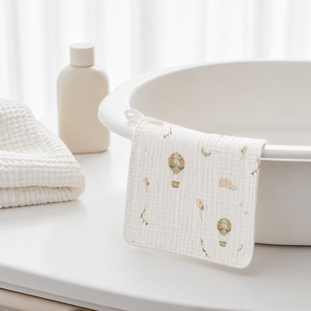 A white muslin baby towel with a delicate hot air balloon pattern draped over the edge of a white bathtub, with a folded textured white towel and a neutral bottle in the background.
