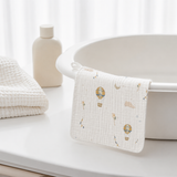 A white muslin baby towel with a delicate hot air balloon pattern draped over the edge of a white bathtub, with a folded textured white towel and a neutral bottle in the background.