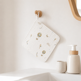 A square white cotton muslin cloth with a delicate hot air balloon and moon pattern, hanging from a wooden peg on a bathroom wall above a sink.