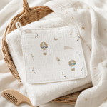 A square muslin baby towel with a hot air balloon and moon print, displayed on top of a folded white terry cloth and a white muslin blanket with gold star prints inside a wicker basket.