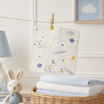 A personalized baby washcloth with a space theme, featuring the name 'LUNARI' and printed with stars, planets, and crescent moons, hanging from a wooden clothespin on a string. The cloth is styled in a nursery setting next to a knitted bunny doll and a basket of folded blankets.