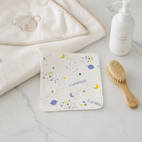 A styled flat-lay of baby bath accessories on a white marble surface, featuring a square muslin towel with a blue and yellow space pattern and the name 'LUNARIS', a folded cream hooded towel with an embroidered bear face, a wooden baby brush, and a white pump bottle labeled 'mild lotion'.