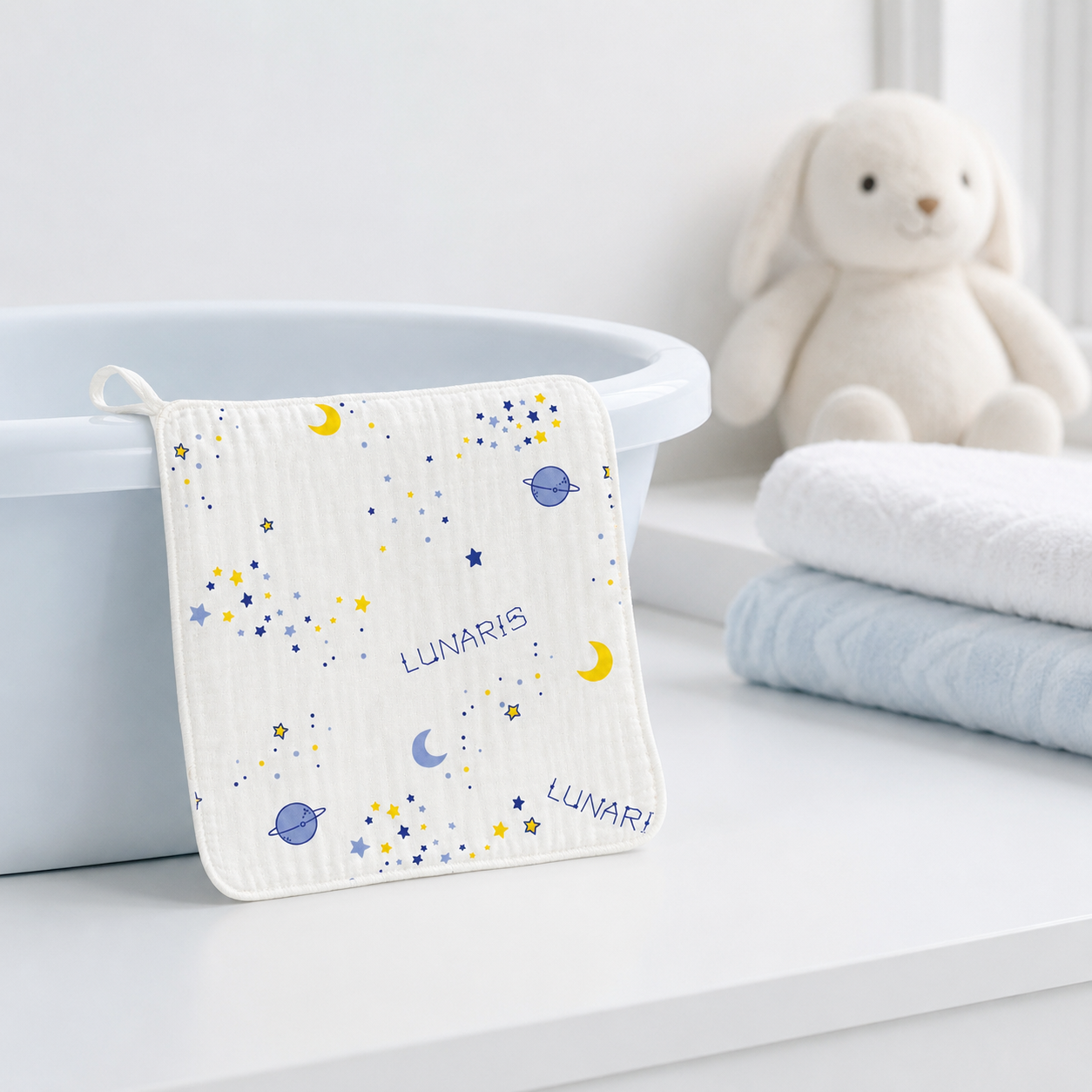 A square white baby washcloth with a blue and yellow space print and the word 'LUNARIS' hangs from a loop next to a white tub, with folded white and light blue towels and a cream bunny toy in the background.