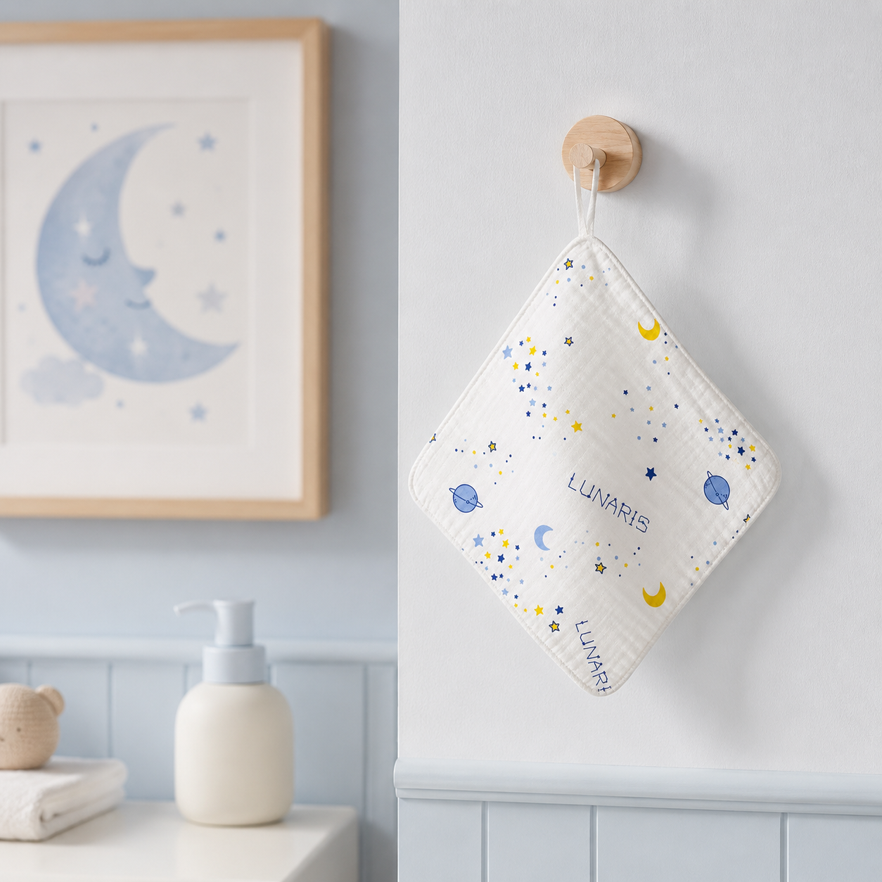 A personalized white quilted cloth pouch with a blue and yellow space print hangs from a wooden peg on a light blue wall in a nursery setting. The pouch is personalized with the name 'LUNARIS' and features a small wooden moon picture on the left.
