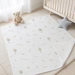 A white quilted baby blanket with a soft pastel print of hot air balloons, clouds, and moons, styled on a cream rug next to a wooden crib, a teddy bear, and a star pillow.
