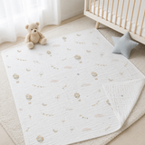 A white quilted baby blanket with a soft pastel print of hot air balloons, clouds, and moons, styled on a cream rug next to a wooden crib, a teddy bear, and a star pillow.