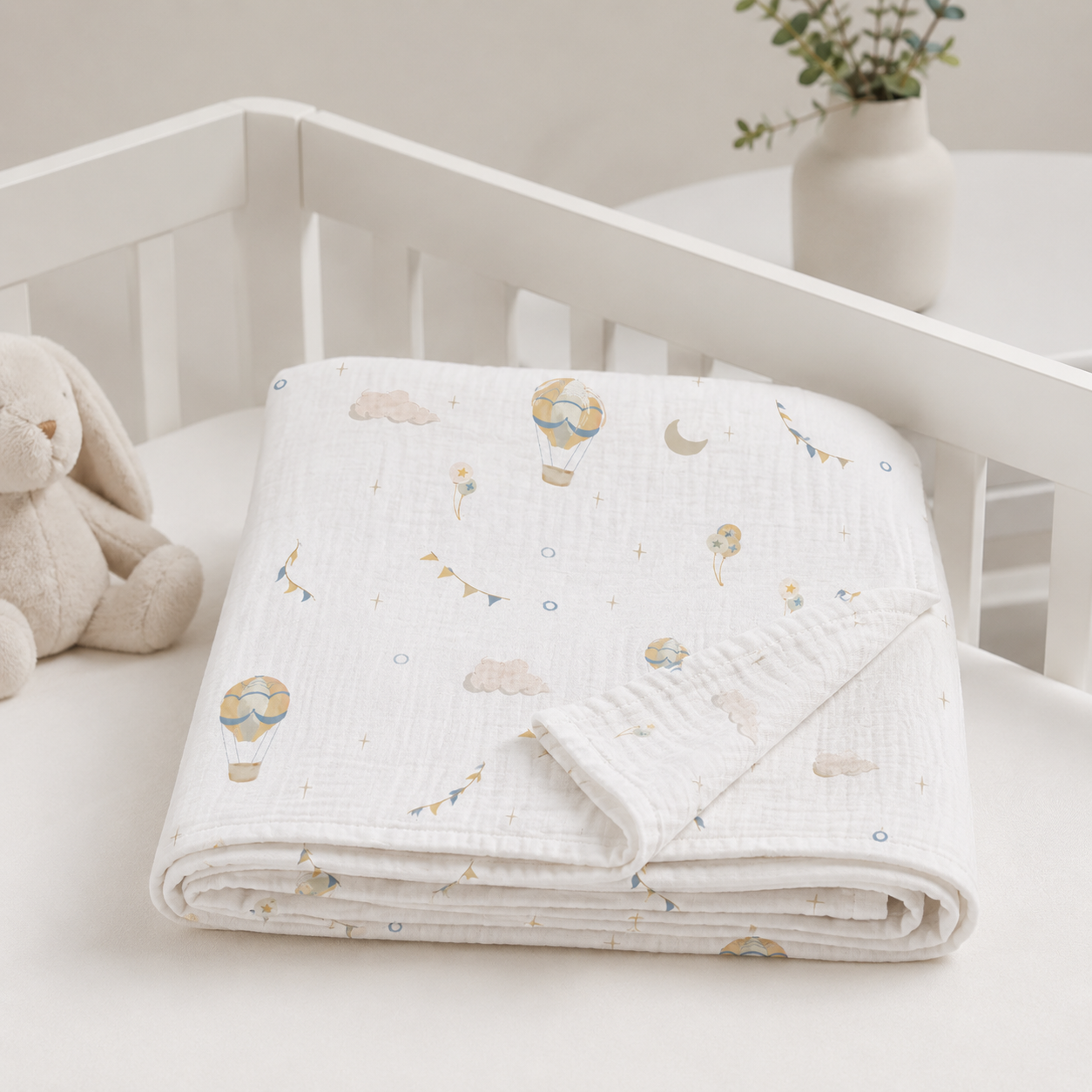 A folded white muslin baby blanket with a soft pastel print of hot air balloons, clouds, and stars, displayed on a white baby crib next to a plush bunny toy.