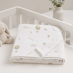 A folded white muslin baby blanket with a soft pastel print of hot air balloons, clouds, and stars, displayed on a white baby crib next to a plush bunny toy.