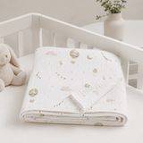 A folded white muslin baby blanket with a soft pastel print of hot air balloons, clouds, and stars, displayed on a white baby crib next to a plush bunny toy.