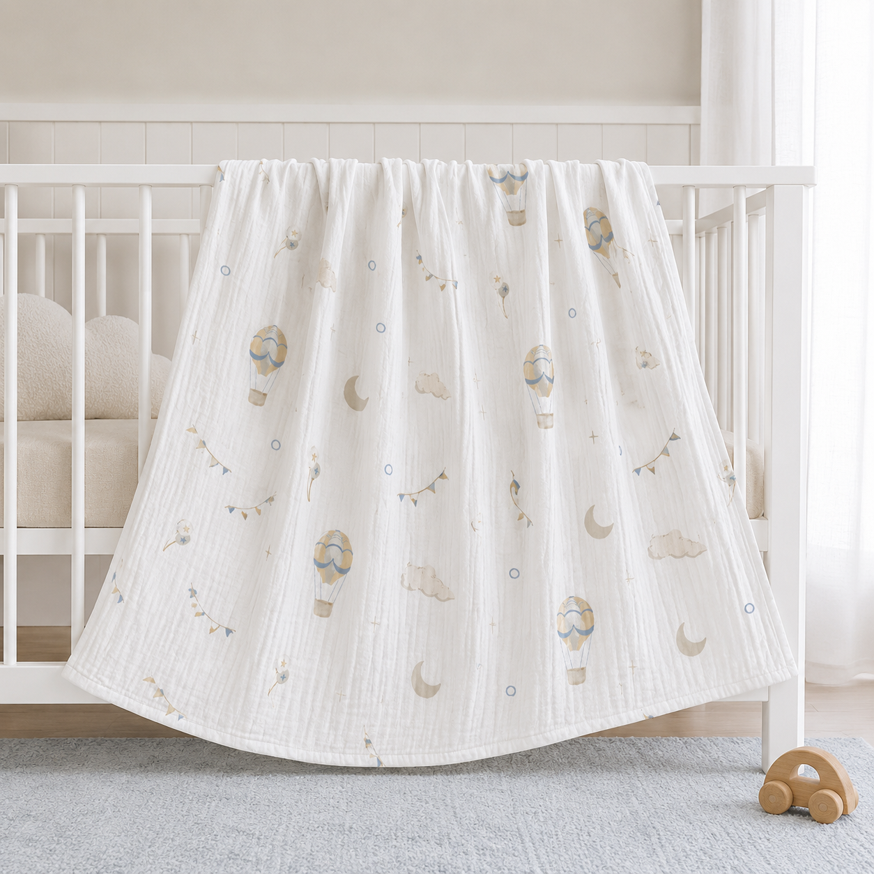 A white muslin baby blanket with a printed pattern of hot air balloons, clouds, and crescent moons, draped over the side of a white wooden baby crib.