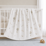 A white muslin baby blanket with a printed pattern of hot air balloons, clouds, and crescent moons, draped over the side of a white wooden baby crib.