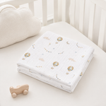 A folded white muslin baby blanket with a soft pastel print of hot air balloons, clouds, and stars, displayed on a white baby crib next to a plush bunny toy.
