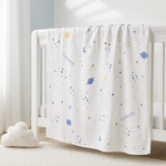 A white muslin baby blanket with a blue and yellow space print, draped over a white wooden crib in a softly lit nursery.
