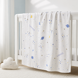 A white muslin baby blanket with a blue and yellow space print, draped over a white wooden crib in a softly lit nursery.