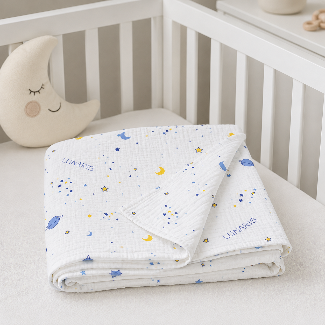 A folded white muslin baby blanket with a blue and yellow star and moon pattern, personalized with the name 'LUNARIS', displayed on a white baby crib next to a crescent moon pillow.
