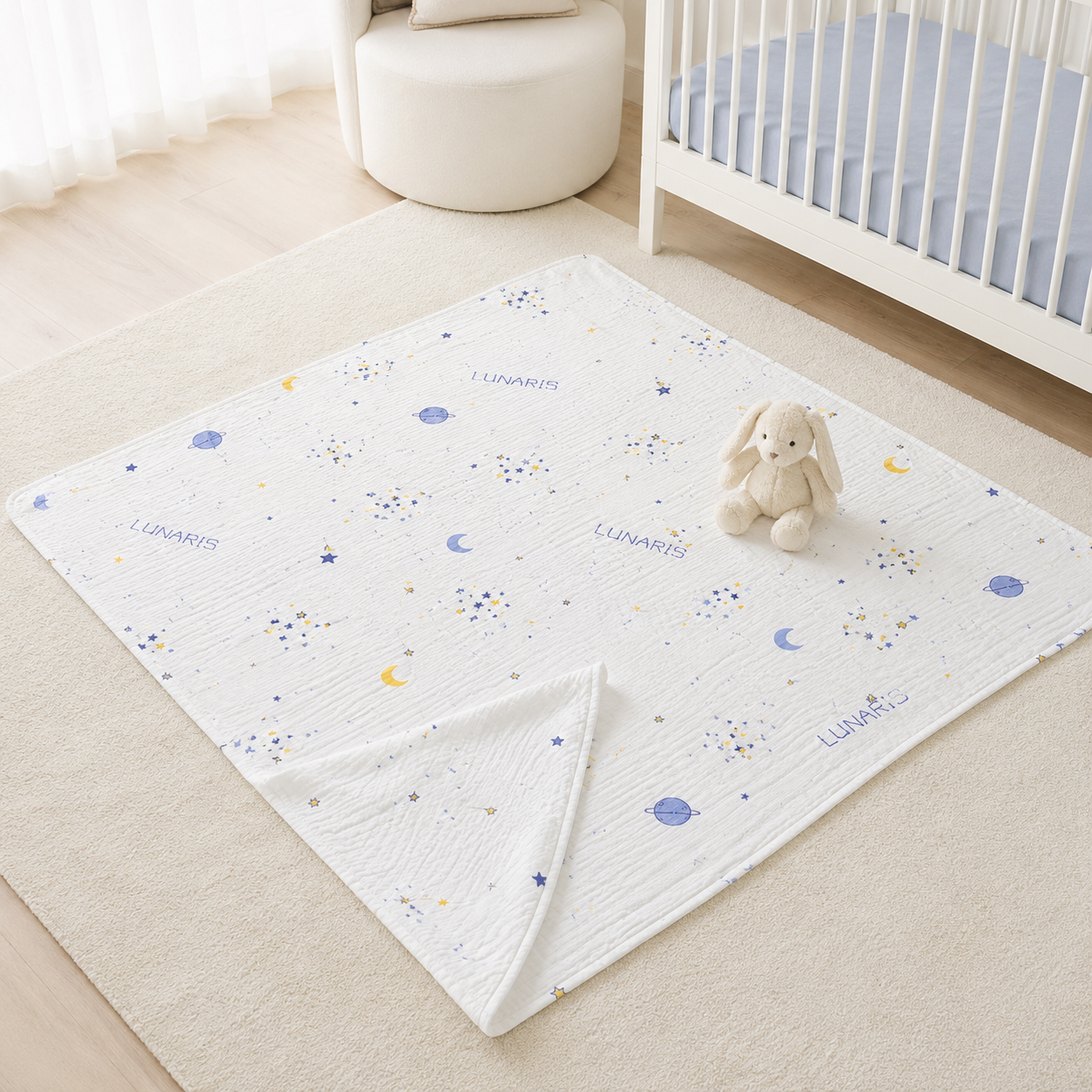 A white muslin baby blanket with a blue and yellow space-themed print of stars, moons, and the words 'LUNARITY' and 'GEMINIS' is styled on a beige rug in a nursery. A small white bunny plush toy sits on the blanket next to a white crib.