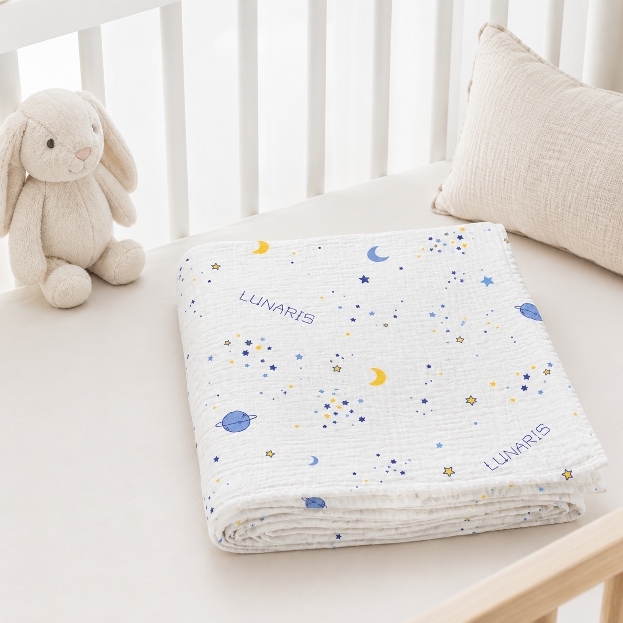 A folded white muslin blanket with a blue and yellow space print, including stars, planets, and crescent moons, and the brand name 'LUNARIS' printed on it. It is styled on a baby bed next to a plush bunny toy and a textured pillow.
