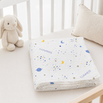 A folded white muslin blanket with a blue and yellow space print, including stars, planets, and crescent moons, and the brand name 'LUNARIS' printed on it. It is styled on a baby bed next to a plush bunny toy and a textured pillow.