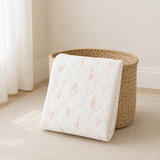 A folded white quilted mattress topper or baby blanket with a soft pink ballet dancer pattern, draped over a woven straw basket on a light beige carpet.