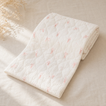 A folded white quilted muslin blanket with a delicate pink bow pattern, styled on a beige linen surface with dried floral accents.
