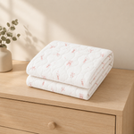 A folded white quilted blanket with a delicate pink bunny pattern sits on a light wood dresser next to a small vase with greenery.