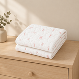 A folded white quilted blanket with a delicate pink bunny pattern sits on a light wood dresser next to a small vase with greenery.