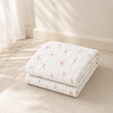 A folded white quilted blanket with a delicate pink bunny pattern, displayed on a textured beige rug in a softly lit room.
