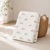 A folded white quilted blanket with a repeating pattern of sleeping cartoon animals in muted green and yellow, styled on a light beige textured rug next to a woven straw basket.