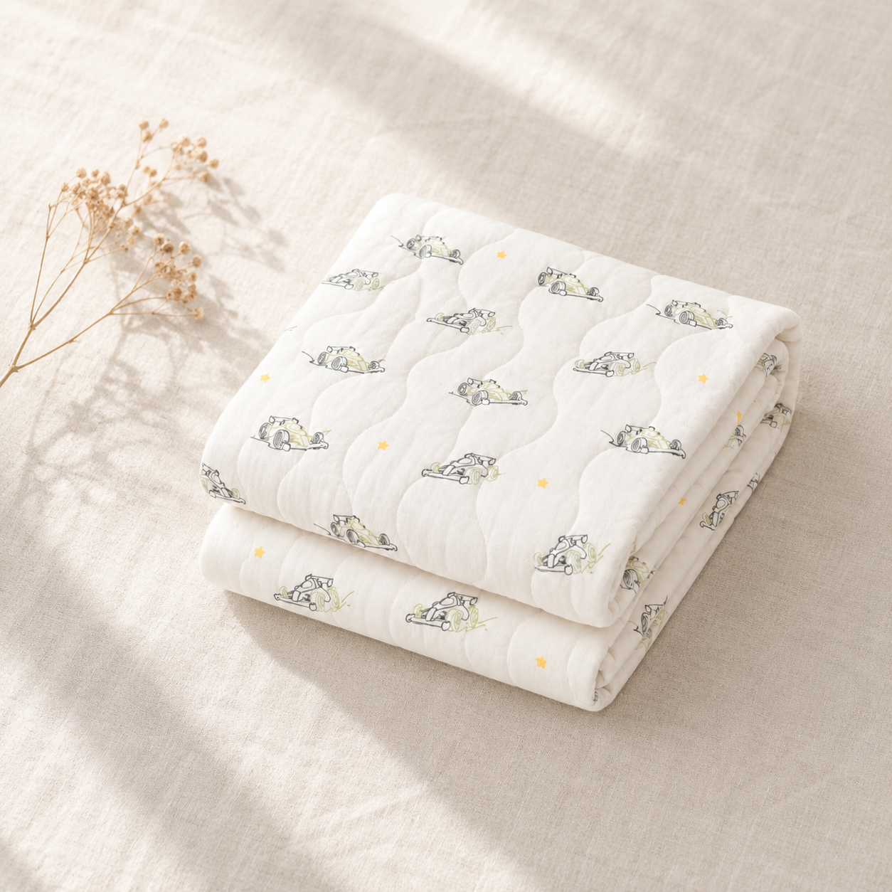 A folded white quilted baby blanket with a cute cartoon cat pattern and small yellow star accents, styled on a beige linen fabric background with dried flowers.