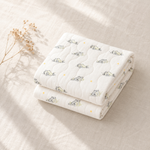 A folded white quilted baby blanket with a cute cartoon cat pattern and small yellow star accents, styled on a beige linen fabric background with dried flowers.