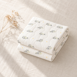 A folded white quilted baby blanket with a cute cartoon cat pattern and small yellow star accents, styled on a beige linen fabric background with dried flowers.