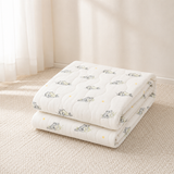 Two folded white quilted baby blankets with a green frog and yellow star pattern, stacked on a textured beige rug in a softly lit room.