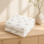 Two folded white quilted blankets with a small bunny print and yellow stars, stacked on a light wood dresser next to a ceramic vase with a plant branch.