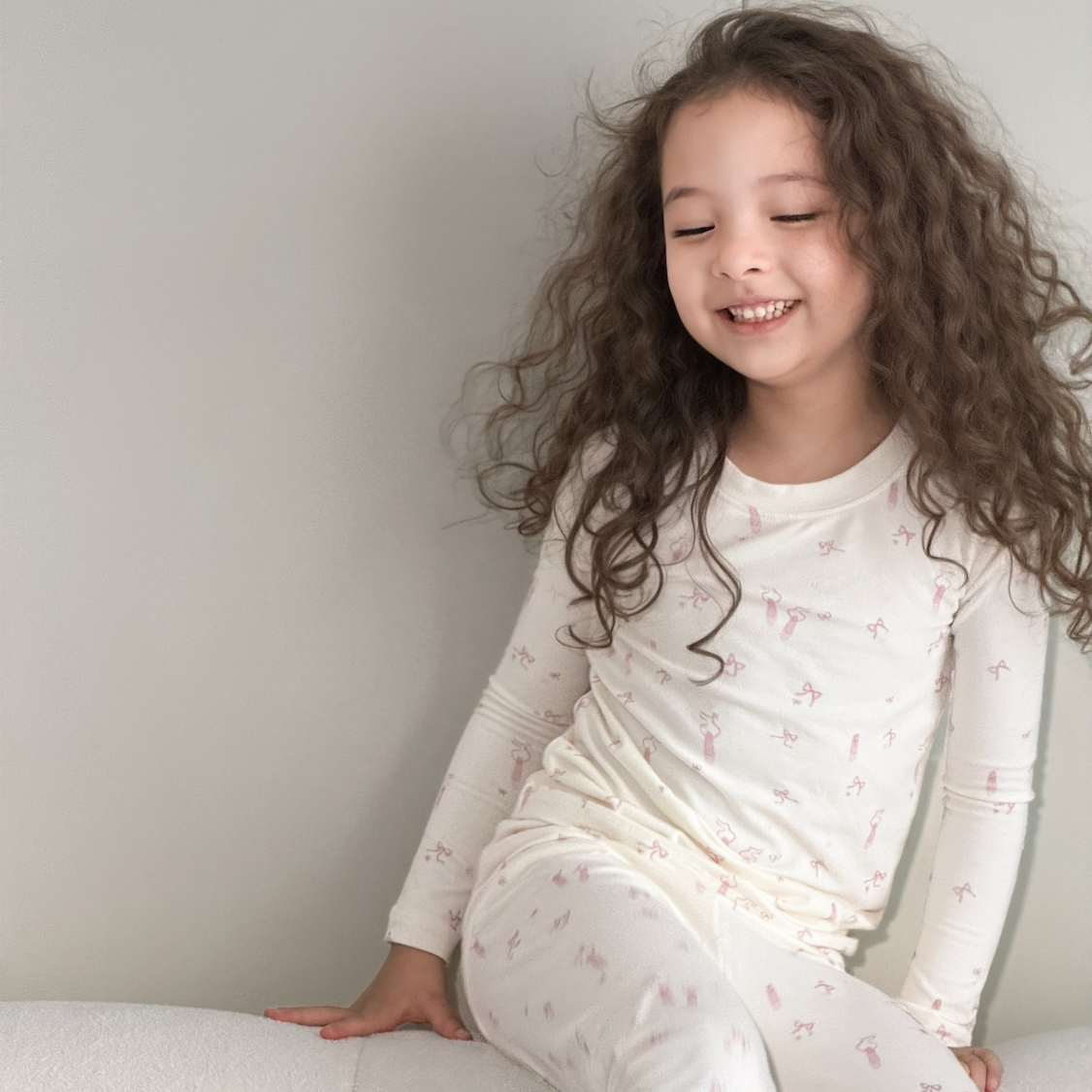 A young girl with long, curly brown hair sits smiling on a light-colored cushion against a plain light gray wall. She is wearing a matching long-sleeve pajama set in cream white with a delicate pink pattern of small butterflies and stars.