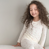 A young girl with long, curly brown hair sits smiling on a light-colored cushion against a plain light gray wall. She is wearing a matching long-sleeve pajama set in cream white with a delicate pink pattern of small butterflies and stars.