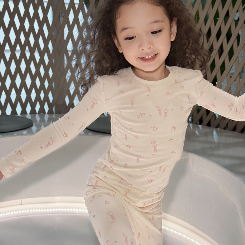 A young girl with curly brown hair wearing a cream-colored long-sleeve pajama set with a pink ballerina pattern, sitting on a white curved play structure.