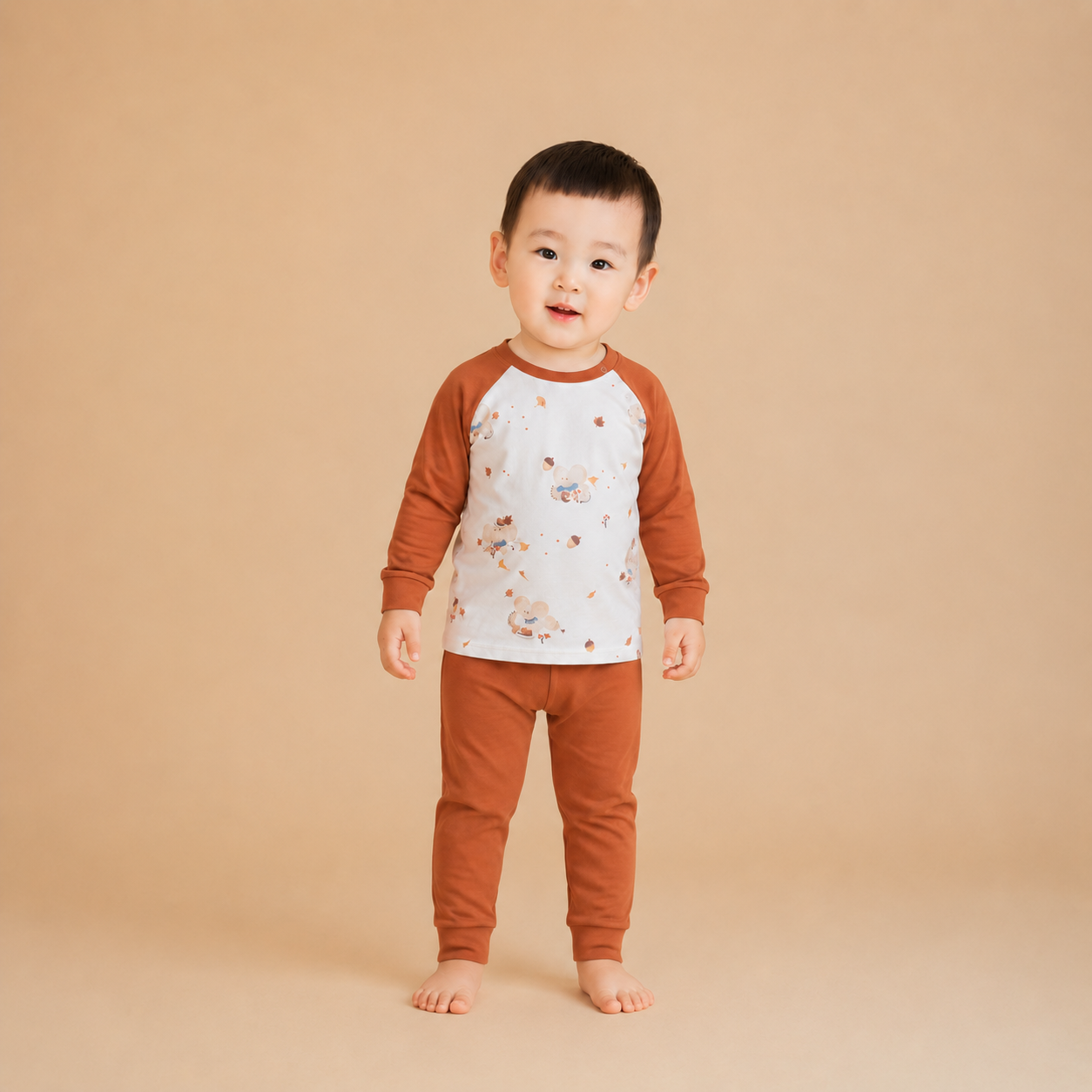 A toddler standing barefoot against a solid beige studio background, wearing a two-piece pajama set with a white body and brown sleeves and pants, decorated with small teddy bear illustrations.