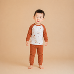 A toddler standing barefoot against a solid beige studio background, wearing a two-piece pajama set with a white body and brown sleeves and pants, decorated with small teddy bear illustrations.