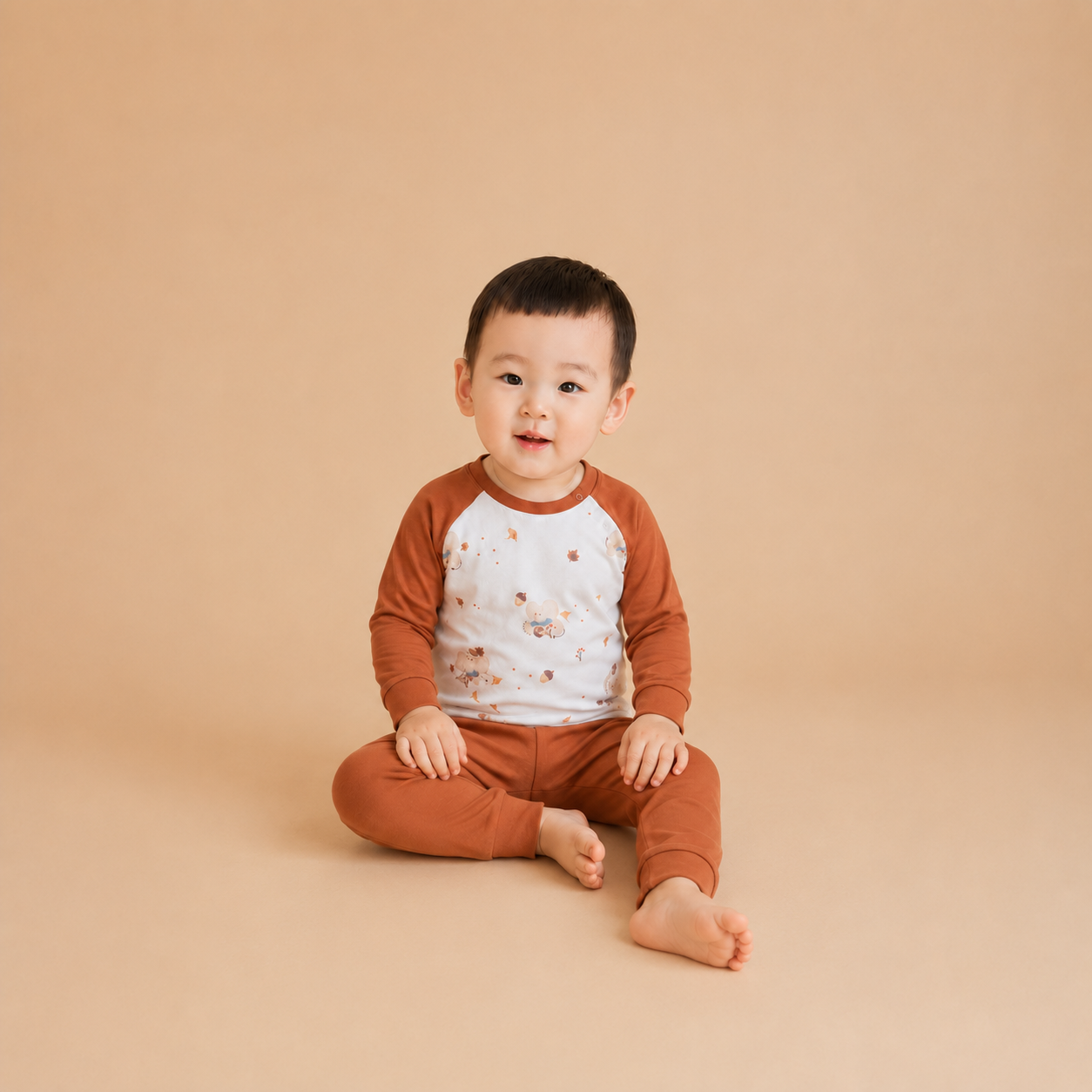 A baby sitting cross-legged on a solid beige studio background, wearing a two-piece raglan pajama set with rust-brown sleeves and pants and a white chest panel with a small floral pattern.