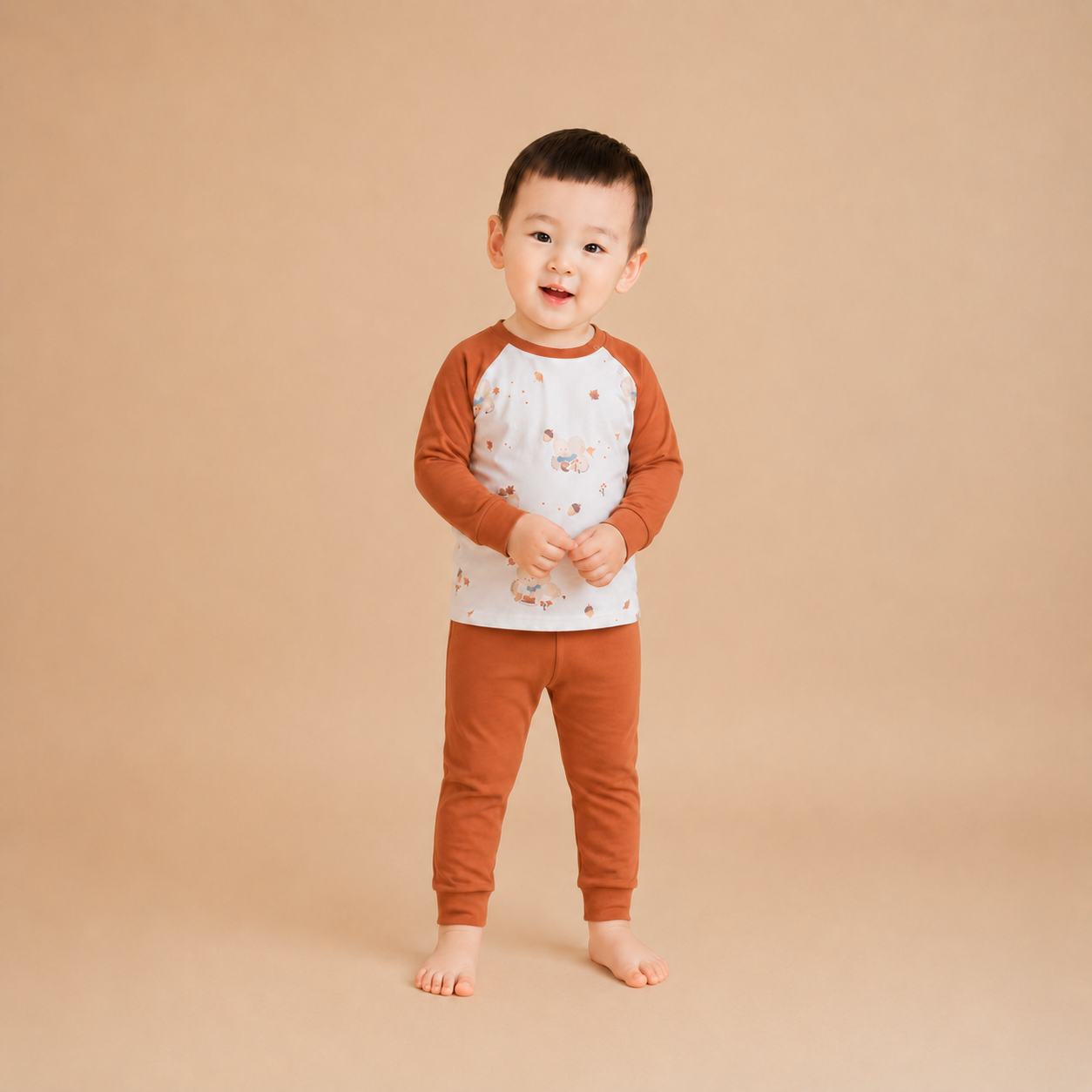 A toddler standing barefoot against a solid beige studio background, wearing a long-sleeve raglan pajama top with a white body and brown sleeves, decorated with small animal illustrations, paired with matching brown pants.
