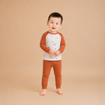 A toddler standing barefoot against a solid beige studio background, wearing a long-sleeve raglan pajama top with a white body and brown sleeves, decorated with small animal illustrations, paired with matching brown pants.
